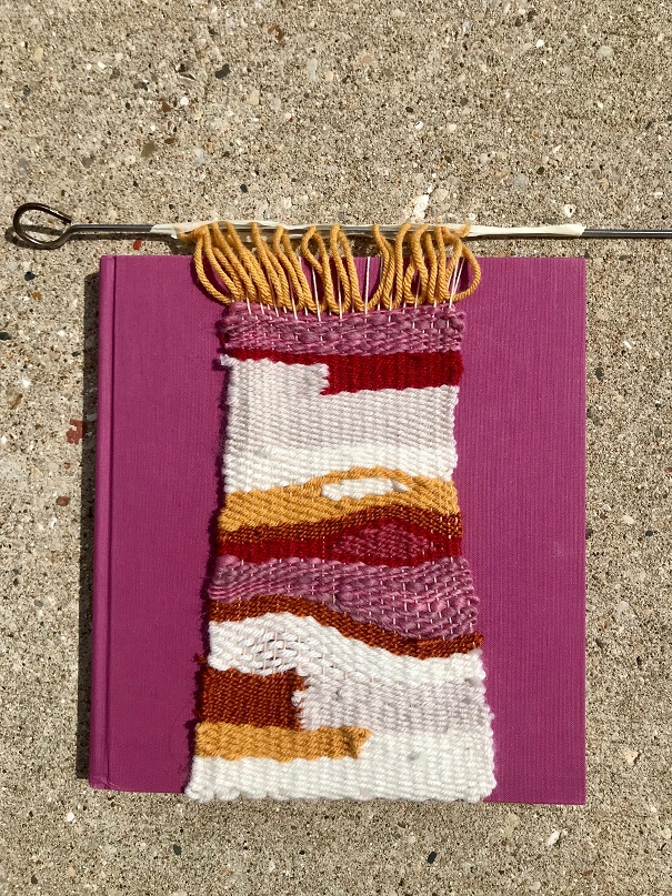 white, cream, pink, yellow, red weaving over pink book cover, pictured on sidewalk