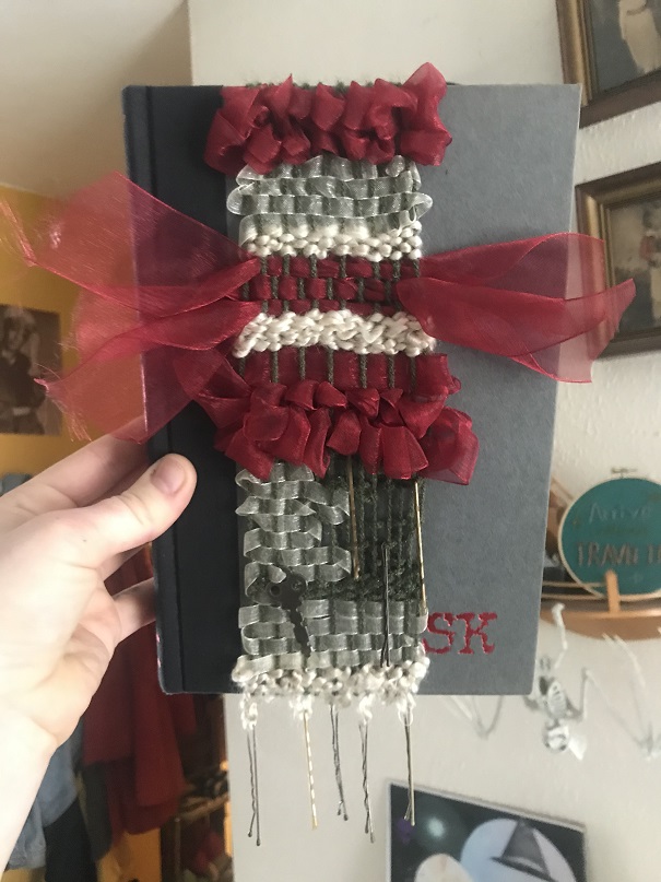 maroon, white, green, gray weaving with bobby pins hanging off the bottom over dark gray book cover