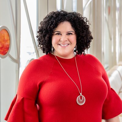 A biracial woman smiling, with dark brown, curly hair, wearing a red sweater and silver necklace.