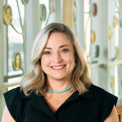 A white woman smiling, with long blonde hair, wearing a black dress and a turquoise necklace.