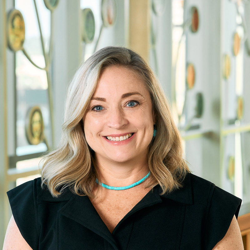 A white woman smiling, with long blonde hair, wearing a black dress and a turquoise necklace.