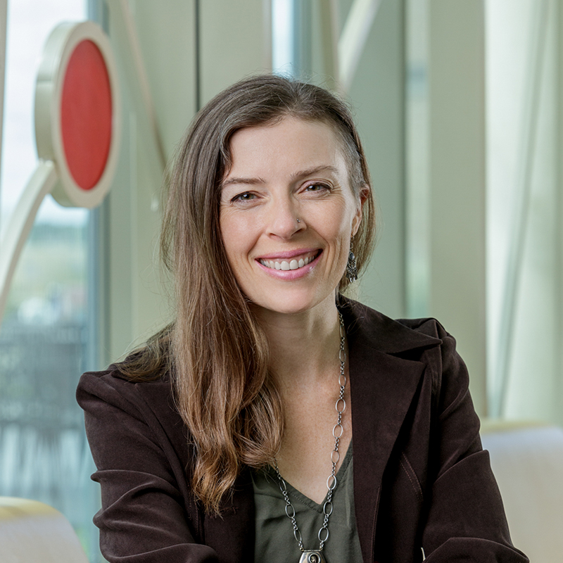 A white woman smiling, with long brown hair, wearing a green dress, a brown blazer and a pendant.