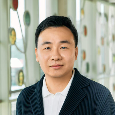 An Asian man smiling, with short black hair, wearing a pleated navy jacket over a white polo shirt.
