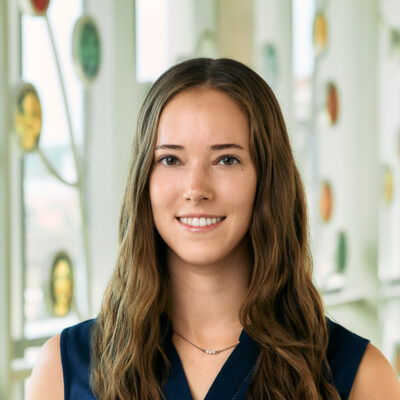 A white woman smiling, with long brown, wavy hair, wearing a navy blue sleeveless top.