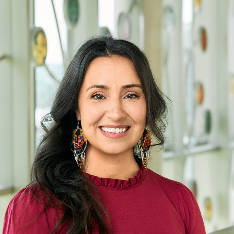 A Middle Eastern woman smiling, with wavy brown hair, wearing a raspberry-colored shirt and large, round, yellow and sparkly earrings.