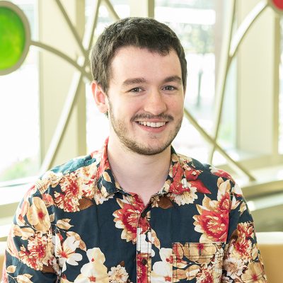 A white male smiling, with short, brown hair and facial hair, wearing a blue shirt with a red and tan flower print.