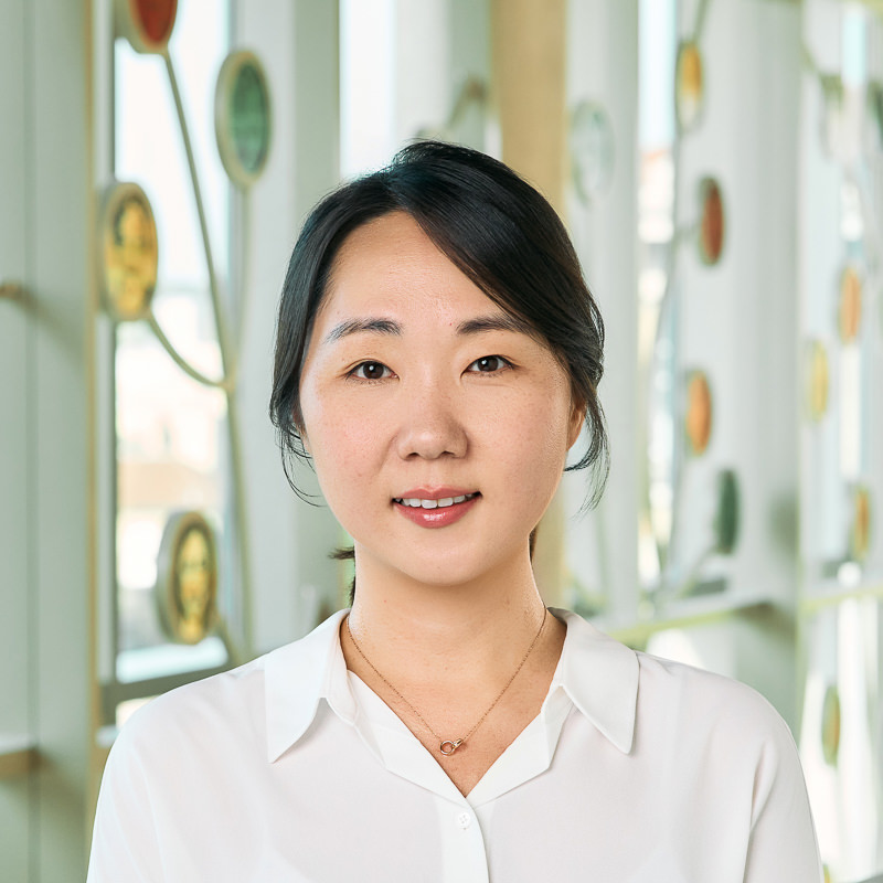 An Asian woman smiling, with dark brown hair in a ponytail, wearing a white blouse with a necklace.