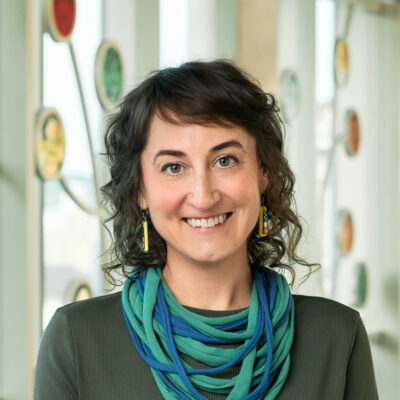 A white woman smiling, with brown, curly hair, wearing a green dress, a layered green and blue cloth necklace, and colorful earrings.