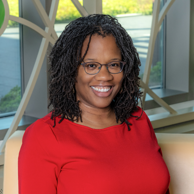 A Black woman, with salt and pepper dreadlocks, wearing glasses and a red dress.
