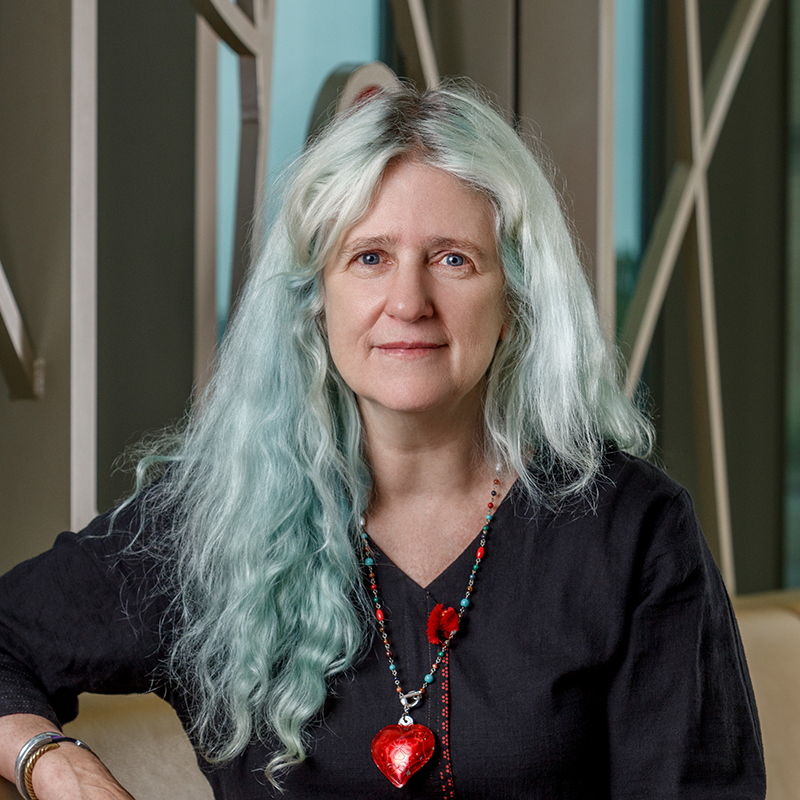 A person with long, blue/green hair, wearing a dark gray top and red necklace.
