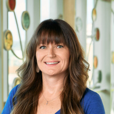 A white woman smiling, with long brown hair, curled with bangs, wearing a blue T-shirt and white earrings.