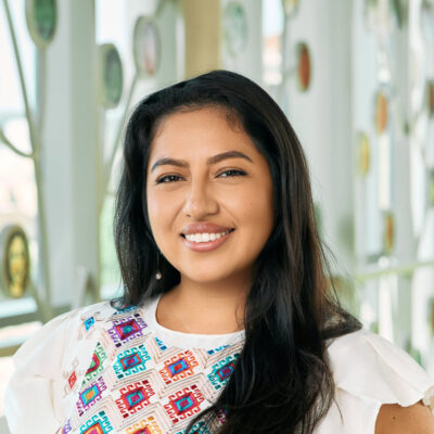 A Latina woman smiling, with long brown hair, wearing a white top with a geometric pattern and dangly pearl earrings.