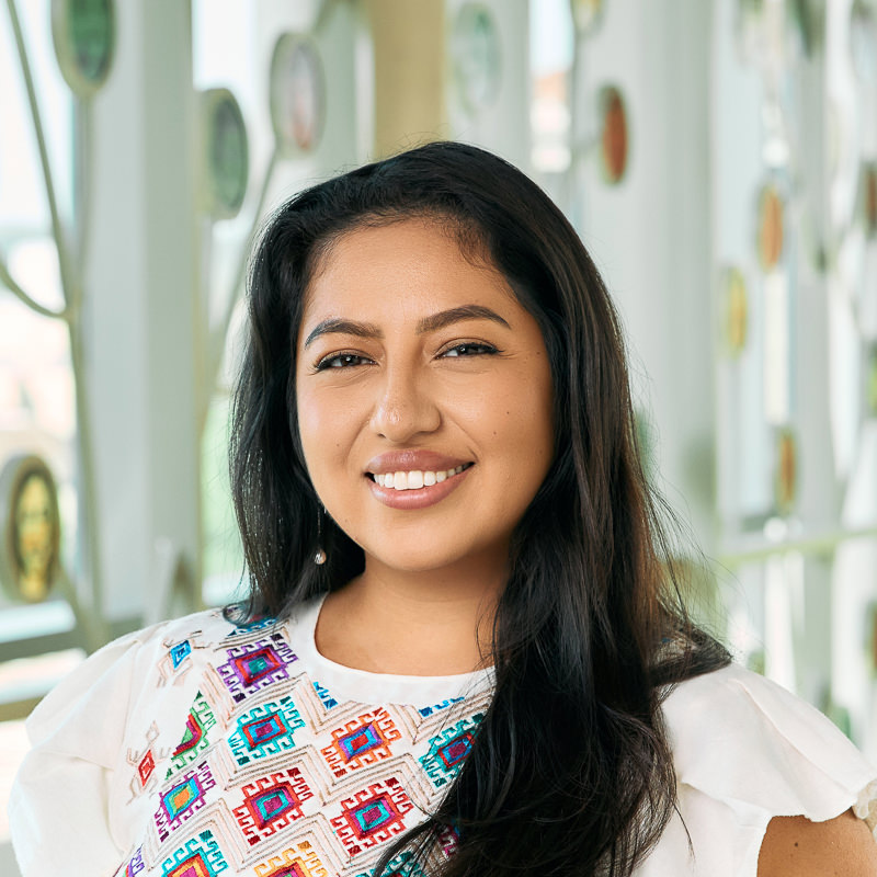 A Latina woman smiling, with long brown hair, wearing a white top with a geometric pattern and dangly pearl earrings.