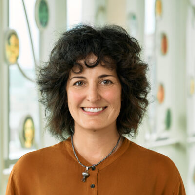 A white woman smiling, with curly, dark brown hair, wearing a copper-colored shirt with a silver necklace.