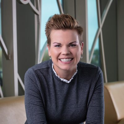 A white woman smiling, with short brown hair, wearing a gray shirt and silver earrings.