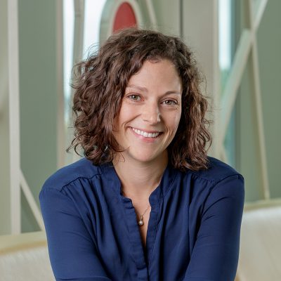 A white woman with brown, curly hair, smiling, wearing a blue dress.
