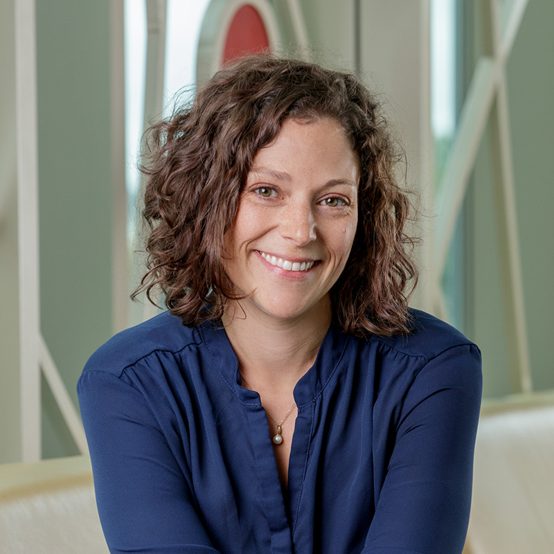 A white woman with brown, curly hair, smiling, wearing a blue dress.