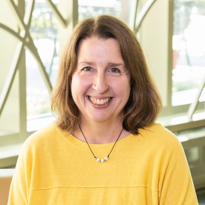 A woman smiling, with shoulder-length brown hair, wearing a yellow shirt and necklace.