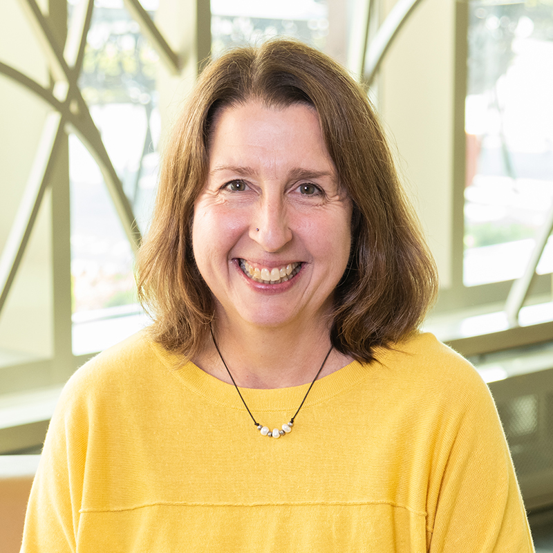 A woman smiling, with shoulder-length brown hair, wearing a yellow shirt and necklace.