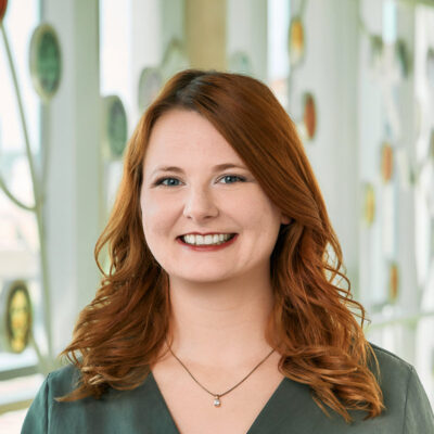 A white woman smiling, with medium-length red hair, wearing a green blouse and gold necklace.
