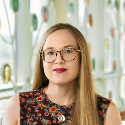 A white woman smiling, with long blonde hair and tortoise shell glasses, wearing a maroon floral print shirt and gold locket.
