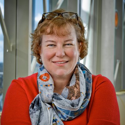 A white woman smiling, with short, red hair, wearing glasses on her head, a patterned scarf around her neck and a red shirt.