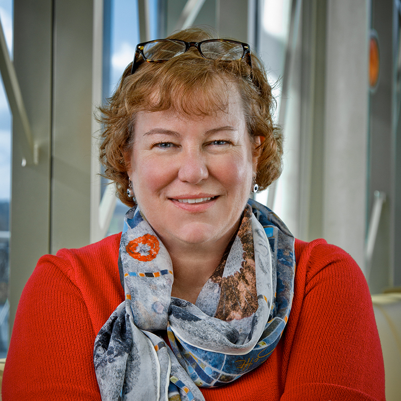 A white woman smiling, with short, red hair, wearing glasses on her head, a patterned scarf around her neck and a red shirt.