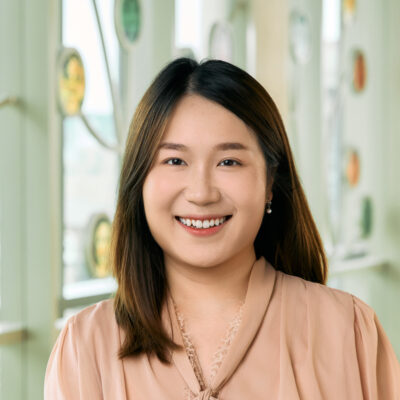 A woman smiling, with short, dark brown hair, wearing a pink blouse and pearl earrings.