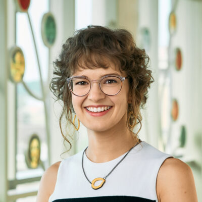 A white woman smiling, with medium-length, brown, curly hair and bangs, wearing glasses, a white shirt, and a yellow necklace.