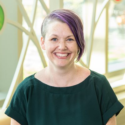 A white woman smiling, with short purple hair, wearing a green shirt.