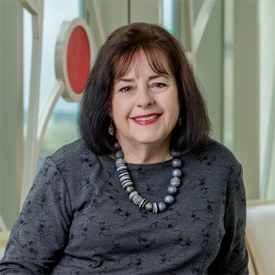 A white woman smiling, with brown hair, wearing a gray top, chunky necklace, and black pants.