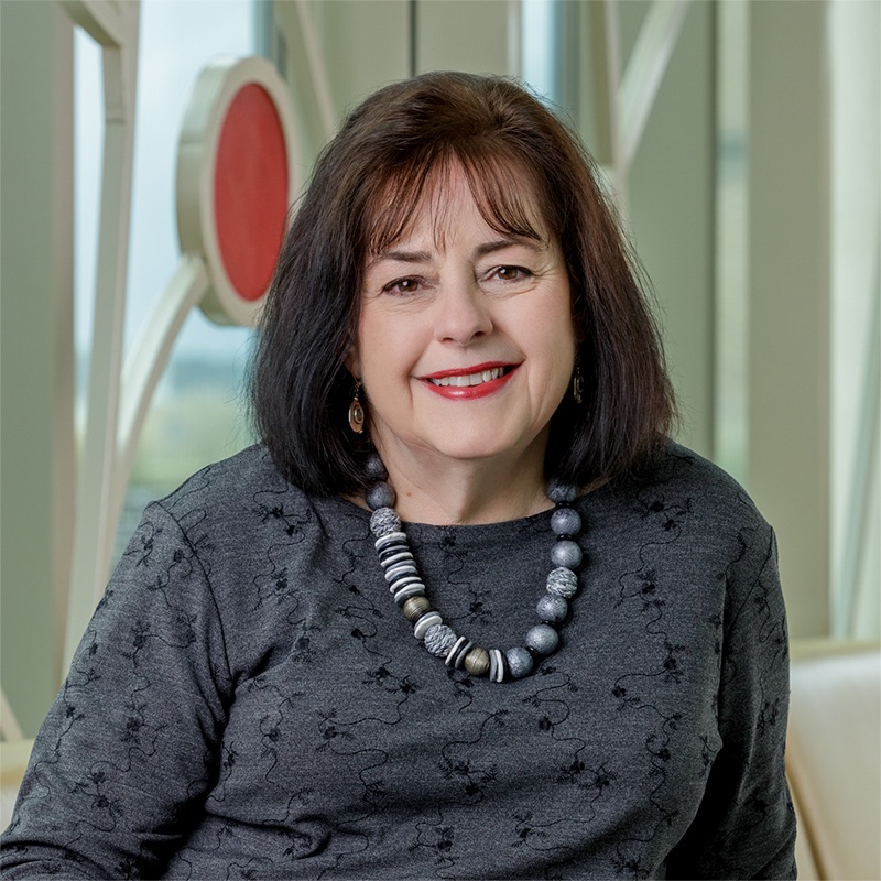 A white woman smiling, with brown hair, wearing a gray top, chunky necklace, and black pants.
