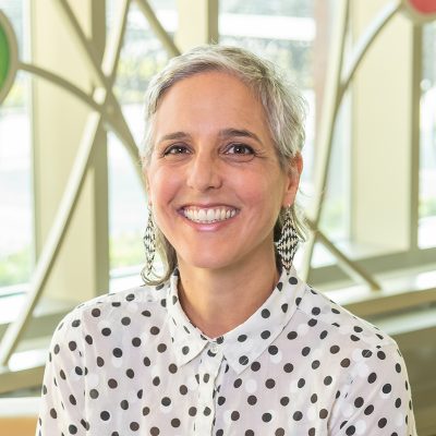 A white woman smiling, with grey hair, wearing graphic-patterned earrings and a polka dot shirt.