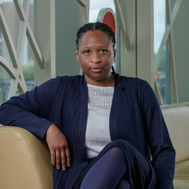 A Black woman sitting legs crossed, wearing a navy blue sweater and skirt and an ivory shirt.