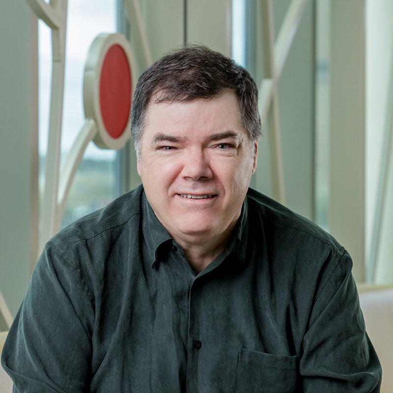 A white man smiling, with dark brown hair, wearing a dark green long-sleeved shirt.