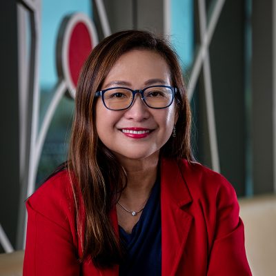 An Asian woman smiling, with shoulder-length dark, straight hair, wearing glasses and a red jacket.