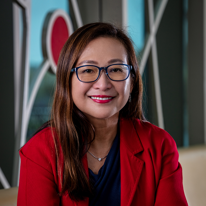 An Asian woman smiling, with shoulder-length dark, straight hair, wearing glasses and a red jacket.