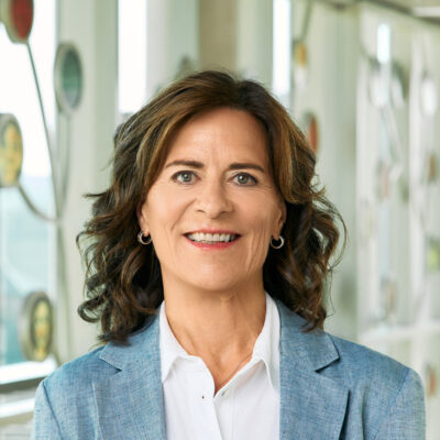 A white woman smiling, with brunette, wavy, shoulder-length hair, wearing a white blouse and chambray blue jacket.