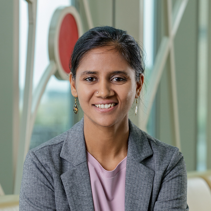 A brown woman with black hair, smiling, wearing a gray jacket and black earrings.