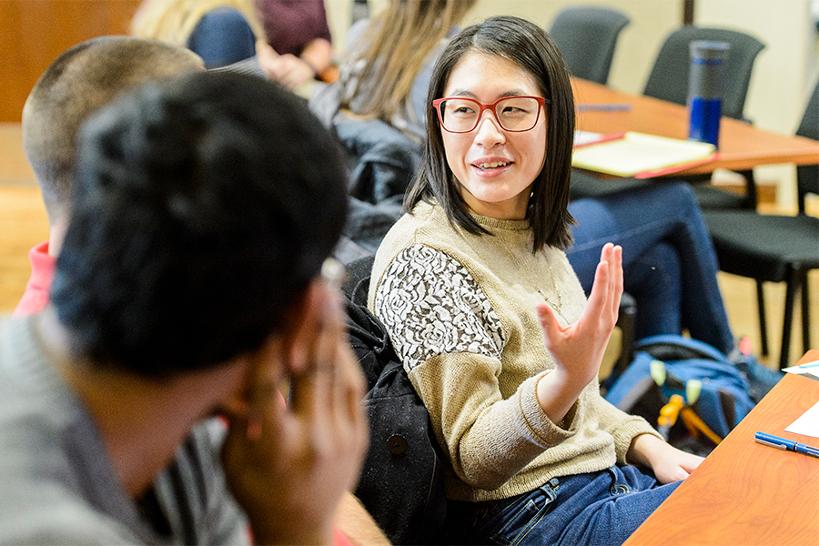 Students sitting at tables talking to each other. One person is in focus, smiling with their hand up as they speak.