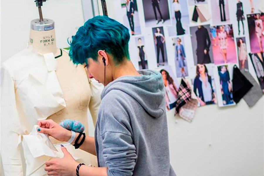 Student pins fabric onto a mannequin, photos of people and fabric samples hang in the background.