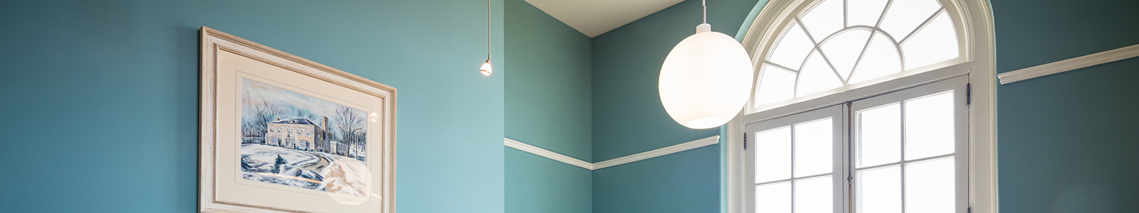 A blue-green room with a framed painting of Nancy Nicholas Hall on the wall, round lamp and an arched window.