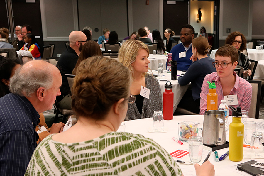 Room full of people discussing around round tables. They're all wearing name tags. Drinks and activities are on the tables.