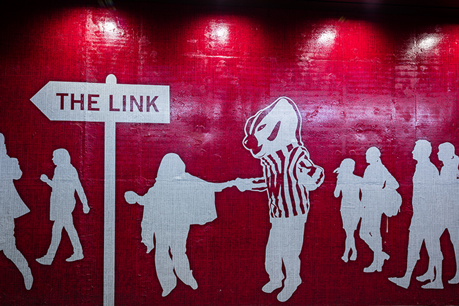 Mural of white silhouettes on a red background. Person holds hands with Bucky Badger while others walk around them.