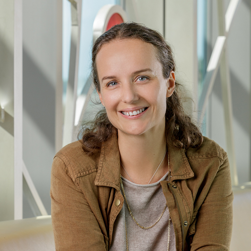A white woman smiling with brown hair, wearing a brown jacket and black pants.