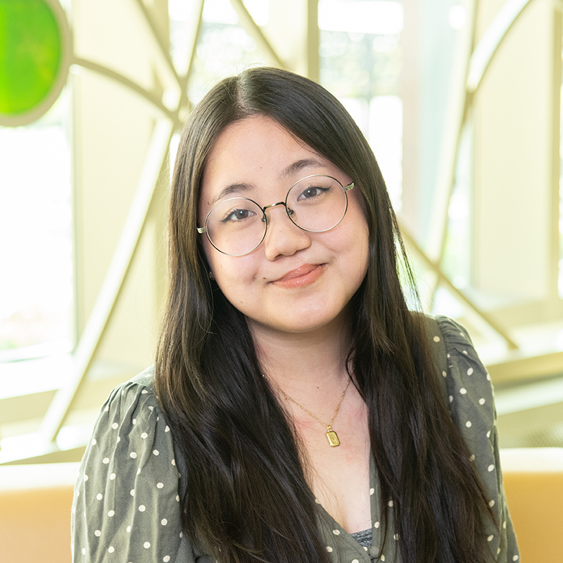 Professional portrait of Jennica Li in front of a decorative window with bright colored glass circles and curved lines.