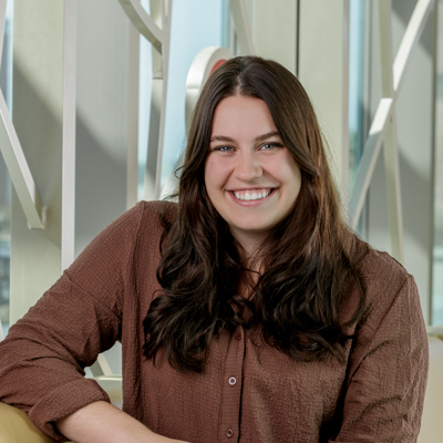 Portrait of Cristin Sutliff, a white woman smiling, with medium-length dark hair, wearing a brown button-up and dark jeans.