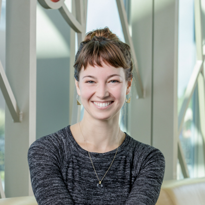 A white woman smiling, wearing a grey dress, with pulled-back hair.