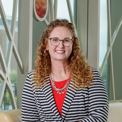 A white woman smiling with glasses, curly red hair, blazer, red blouse, and a gold necklace.