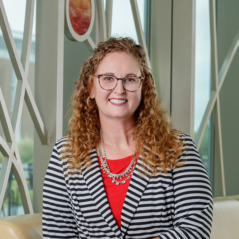 A white woman smiling with glasses, curly red hair, blazer, red blouse, and a gold necklace.